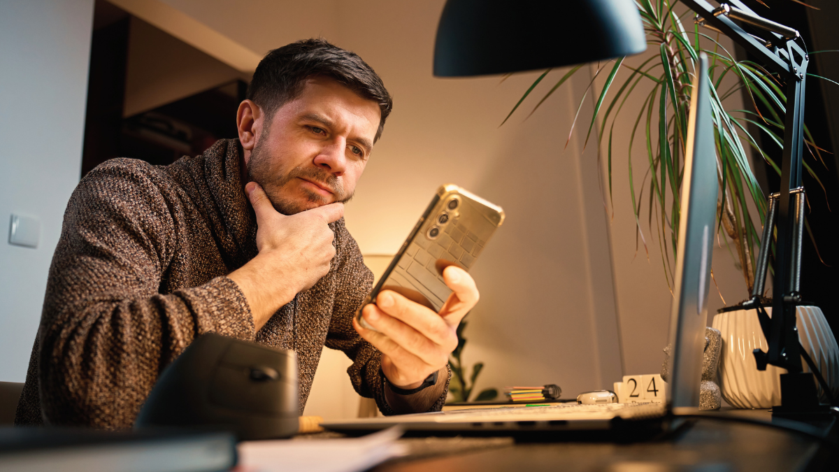 Thoughtful man sitting at a desk, looking at his phone with a skeptical expression, suggesting disengagement with a digital experience.